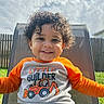 toddler, child, smiling, curly_hair, outdoor, chair, sweater, orange, white, mesh_chair, fence, grass, sunny, cloudy_sky, portrait, happy, casual_clothing, young_child, daylight, backyard