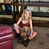 blonde_hair, boots, carpet, casual_clothing, child, companion, couch, decor, dog, furniture, indoor, leather_chair, living_room, pet, relaxing, shelf, sitting, smiling, television, wooden_table