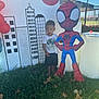 child, boy, grass, spiderman, cutout, cupcakes, balloons, outdoor, fence, cityscape, backdrop, shirt, shorts, barefoot, sunlight, shadow, party, decorations, trees, daytime