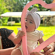 Iris participe au concours pour gagner de l'argent avec cette photo : baby, headband, balloon, pink_balloon, heart_shape, child, hands, outdoor, greenery, garden, table, white_dress, summer, cute, playful, infant, person, closeup, soft_light, family