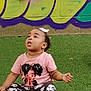 toddler, child, sitting, curious, pink_shirt, cartoon_print, floral_pants, green_turf, graffiti, wall, indoor, casual_clothing, young_child, portrait, cute, looking_up, short_hair, bow, playful, daylight