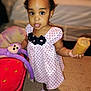 toddler, child, girl, polka_dot_dress, stuffed_animal, toy, purple_monkey, indoor, floor, socks, snack, curious, standing, young_child, person, cute, playful, expression, home, lighting