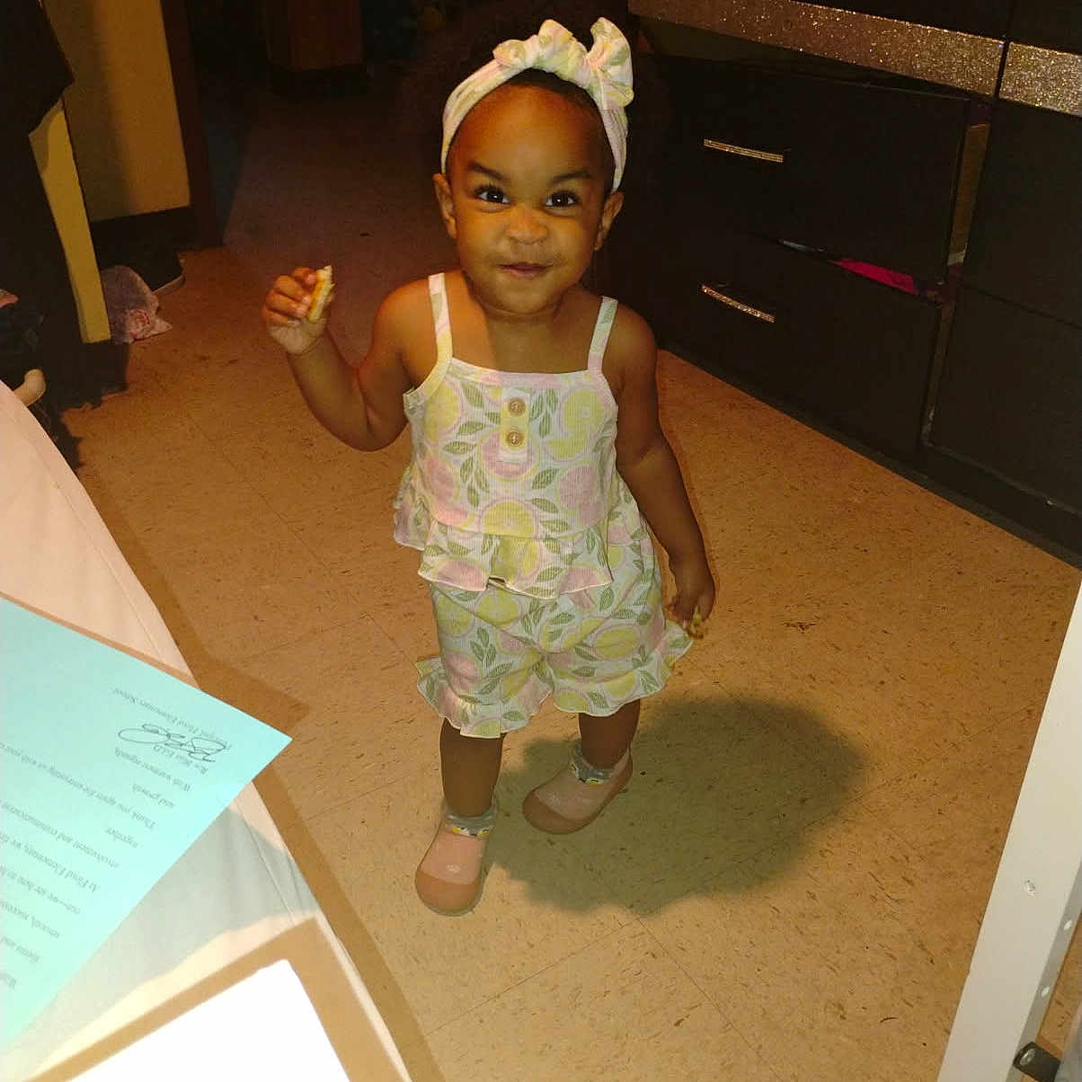 Amora joined the competition — help win amazing prizes! cabinet, child, cute, drawer, expression, floor, floor_tile, furniture, happy, headband, holding_food, indoor, patterned_clothing, person, room, shadow, shoes, smile, standing, toddler