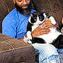 animal, beard, black_and_white_cat, blue_shirt, casual_clothing, cat, couch, expression, facial_hair, feline, hands, holding, home, indoor, jeans, man, pet, relaxed, ripped_jeans, seated