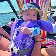 Tommy is registered to the contest to win money with this photo: baby, life_jacket, hat, boat, water, towel, person, sunlight, outdoor, summer, child, holding, smile, clothing, blue_sky, nature, recreation, vacation, fun, family