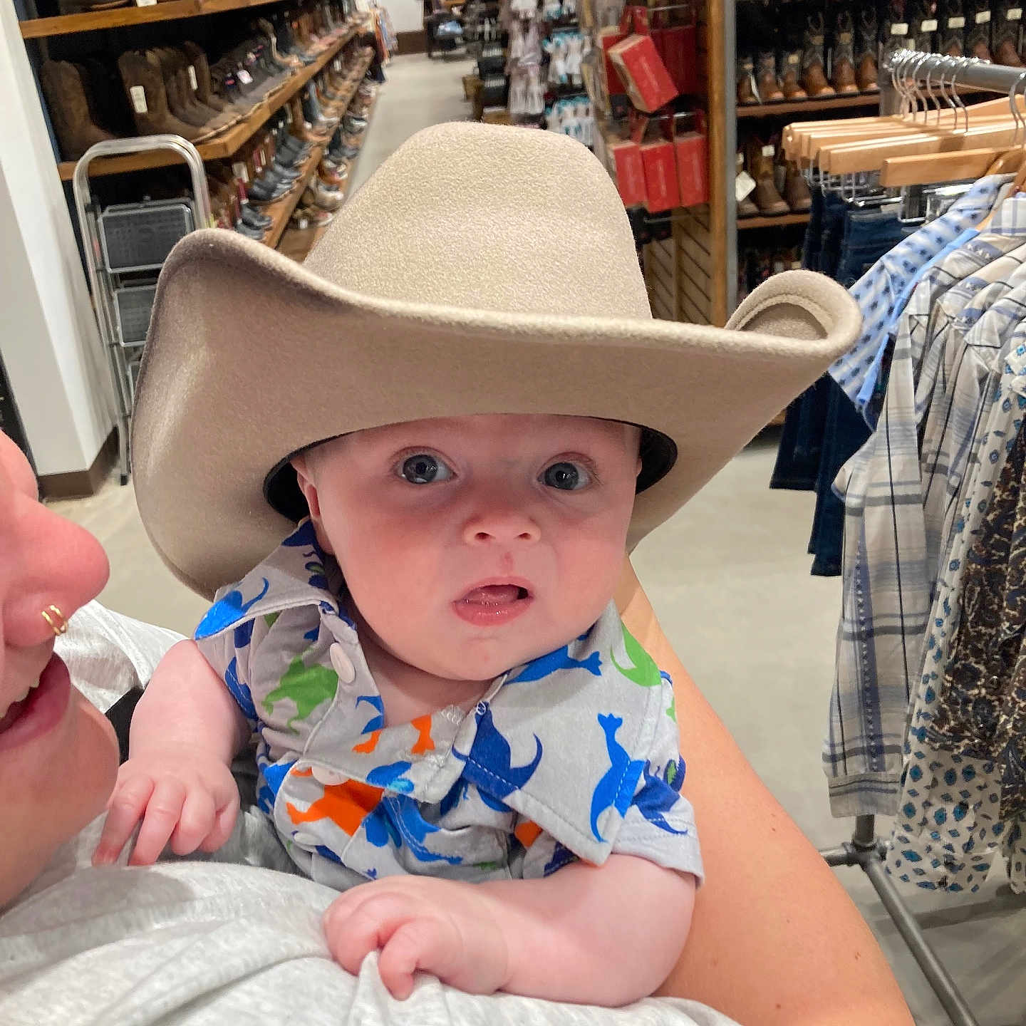 Tommy is registered to the contest to win money with this photo: adult, baby, boots, child, clothing, cowboy_hat, curious, dinosaur_print, expression, face, holding, indoor, person, plaid, rack, shelf, shirt, shopping, store, wide_eyes