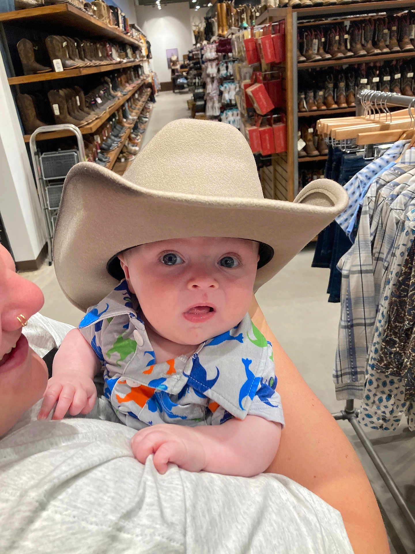 Tommy is registered to the contest to win money with this photo: baby, cowboy_hat, child, person, clothing, store, boots, shelf, rack, shopping, adult, holding, plaid, shirt, dinosaur_print, curious, indoor, face, wide_eyes, expression