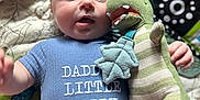 Tommy is registered to the contest to win money with this photo: baby, infant, onesie, text, toy, plush_toy, dinosaur, blue_clothing, blanket, pattern, child, cute, lying_down, indoor, soft_toys, colorful, hand, face, expression, comfort