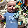 baby, infant, onesie, text, toy, plush_toy, dinosaur, blue_clothing, blanket, pattern, child, cute, lying_down, indoor, soft_toys, colorful, hand, face, expression, comfort