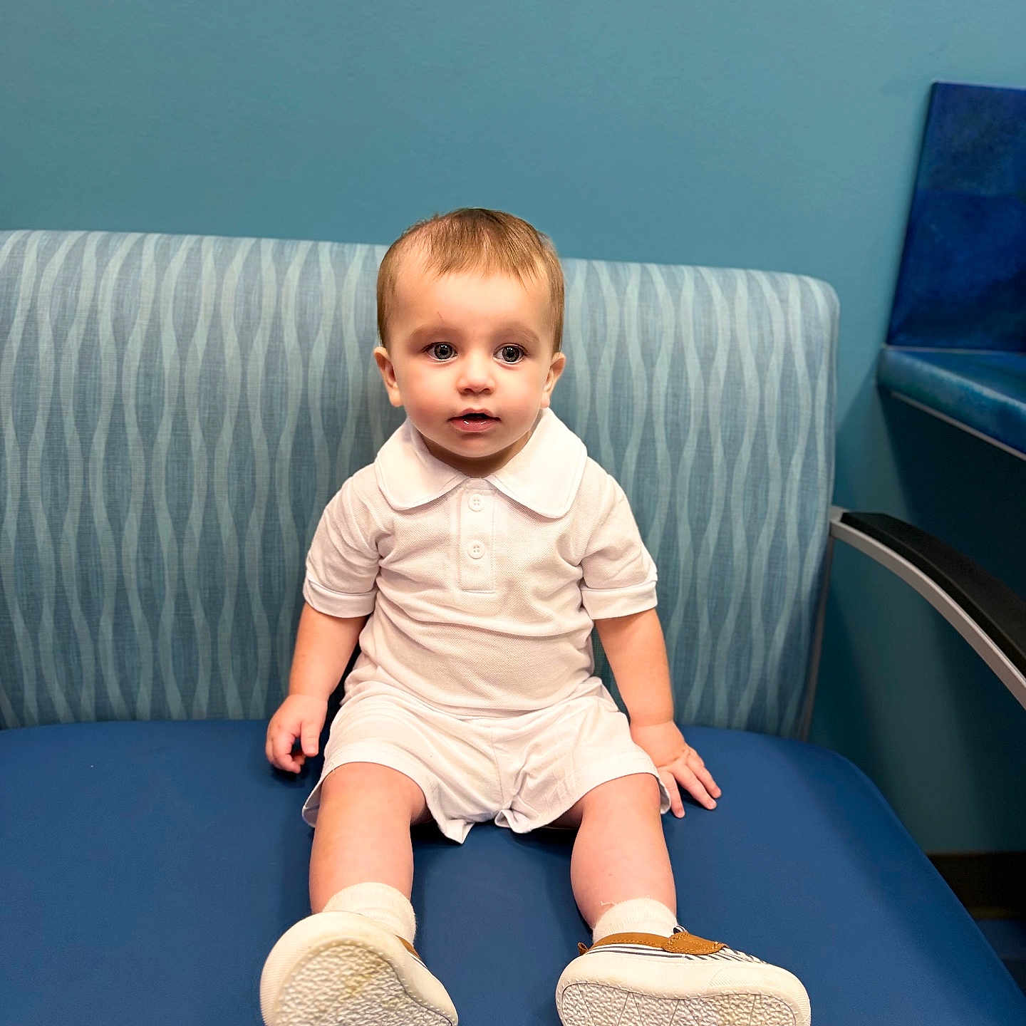 Jdee is registered to the contest to win money with this photo: toddler, child, baby, white_clothing, shorts, polo_shirt, sneakers, socks, blue_bench, patterned_backrest, indoor, seated, curious_expression, young_child, furniture, blue_wall, casual, portrait, person, cute