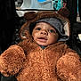 baby, child, toddler, fuzzy_clothing, bear_costume, hood, ears, stroller, seatbelt, mesh_background, cute, portrait, smiling, indoors, brown_clothing, face, eyes, mouth, jacket, warm