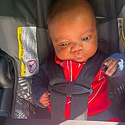 Phillip is registered to the contest to win money with this photo: baby, car_seat, child, infant, seatbelt, red_clothing, black_straps, safety, infant_car_seat, cute, indoors, young_child, face, hands, sunlight, seat, straps, person, child_safety, closeup