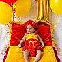 baby, infant, red_clothing, yellow_clothing, pillow, balloons, number_balloon, celebration, birthday, red_balloon, yellow_balloon, plush, cute, portrait, child, festive, soft_texture, smile, hat, lying_down