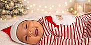 Noah is registered to the contest to win money with this photo: baby, infant, smiling, face, red_hat, striped_clothing, christmas_theme, holiday, festive, soft_surface, portrait, indoors, decorations, lights, tree, happy, cute, infant_clothing, cozy, closeup