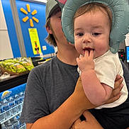 Madelynn is registered to the contest to win money with this photo: baby, child, person, holding, elephant_hat, inflatable, store, indoor, shopping, shelf, water_bottles, gray_shirt, curious, hand, face, headwear, casual_clothing, young, fingers_in_mouth, retail