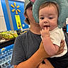 baby, child, person, holding, elephant_hat, inflatable, store, indoor, shopping, shelf, water_bottles, gray_shirt, curious, hand, face, headwear, casual_clothing, young, fingers_in_mouth, retail