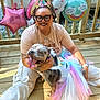 balloons, casual_clothing, celebration, cloud_balloon, costume, dog, glasses, happy, headband, outdoor, person, pet, rainbow, smiling, star_balloon, sunlight, tattoo, tutu, unicorn_costume, wooden_deck