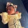 Zahara joined the competition — help win amazing prizes! baby, infant, headband, yellow_clothing, lying_down, curious_expression, face, hand, dark_background, text_on_clothing, closeup, soft_lighting, newborn, cute, portrait, child, indoors, baby_girl, fashion, adorable