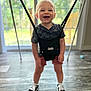 baby_jumper, black_shirt, child, clothing, daylight, floor, fun, happy, home, indoor, person, playful, shoe, shorts, smiling, sneaker, standing, toddler, window, young_child