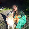 woman, dog, smiling, night, outdoor, green_dress, pet, happy, grass, gravel, car, collar, long_hair, shadow, face, licking, crouching, nature, friendship, light
