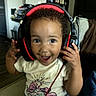 child, toddler, portrait, smiling, happy, headphones, gaming_headset, curly_hair, indoor, living_room, seated, shirt, denim, skirt, hands, camera, close_up, joyful, home, music