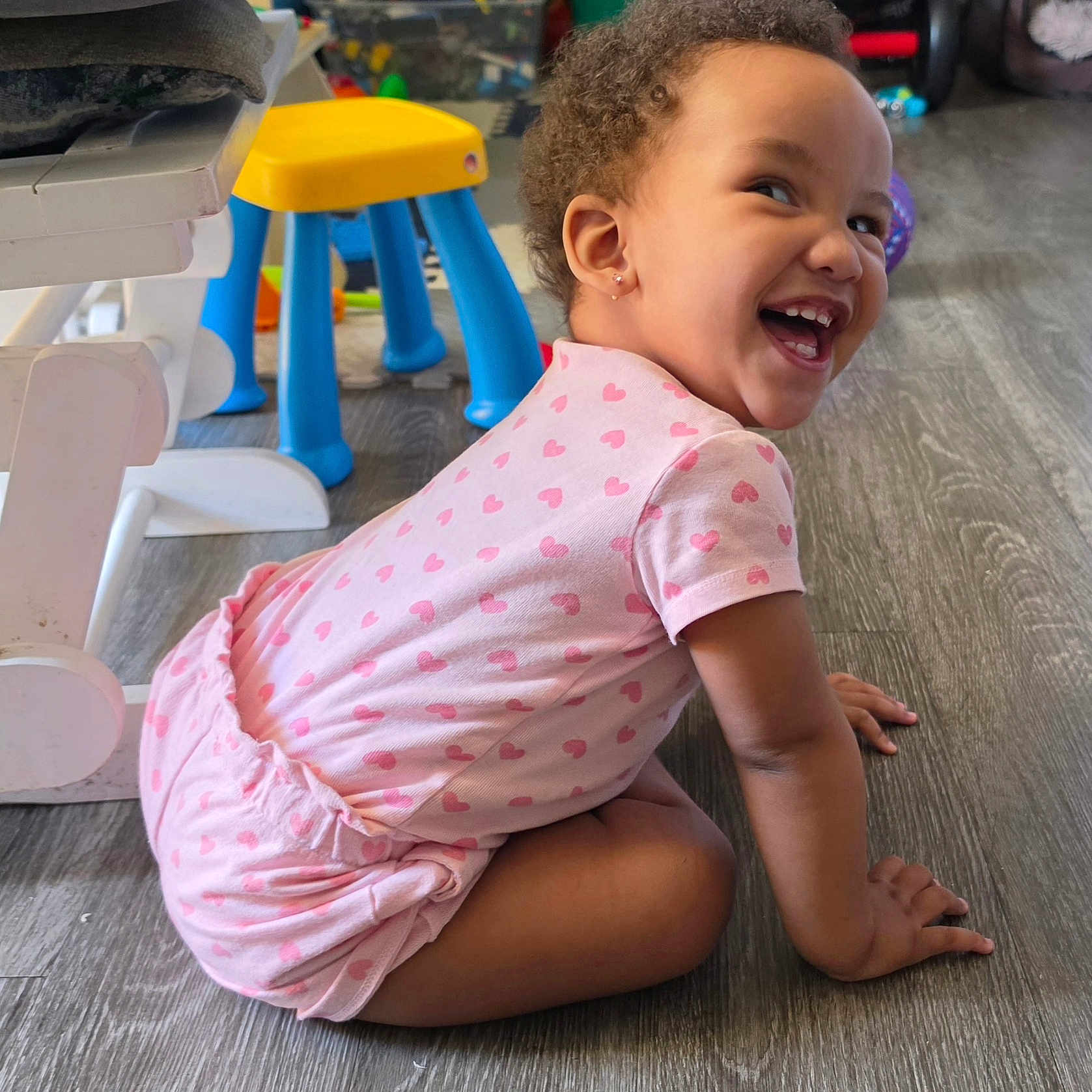 Aaliyah is registered to the contest to win money with this photo: child, toddler, girl, smiling, laughing, portrait, indoor, playroom, pink_clothing, heart_pattern, curly_hair, earrings, sitting, floor, stool, toys, high_chair, wood_floor, happy, candid
