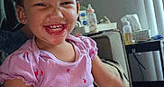 Aaliyah joined the competition — help win amazing prizes! child, girl, toddler, smiling, laughing, portrait, indoor, seated, pink_shirt, red_pants, curly_hair, happy, face, teeth, chair, table, bottles, tissues, household, photography