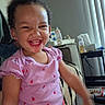 child, girl, toddler, smiling, laughing, portrait, indoor, seated, pink_shirt, red_pants, curly_hair, happy, face, teeth, chair, table, bottles, tissues, household, photography