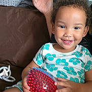 Aaliyah joined the competition — help win amazing prizes! child, toddler, smiling, portrait, indoor, couch, seated, toy, ball, red, blue, patterned_shirt, curly_hair, earrings, hands, playful, happy, home, cushion, photograph
