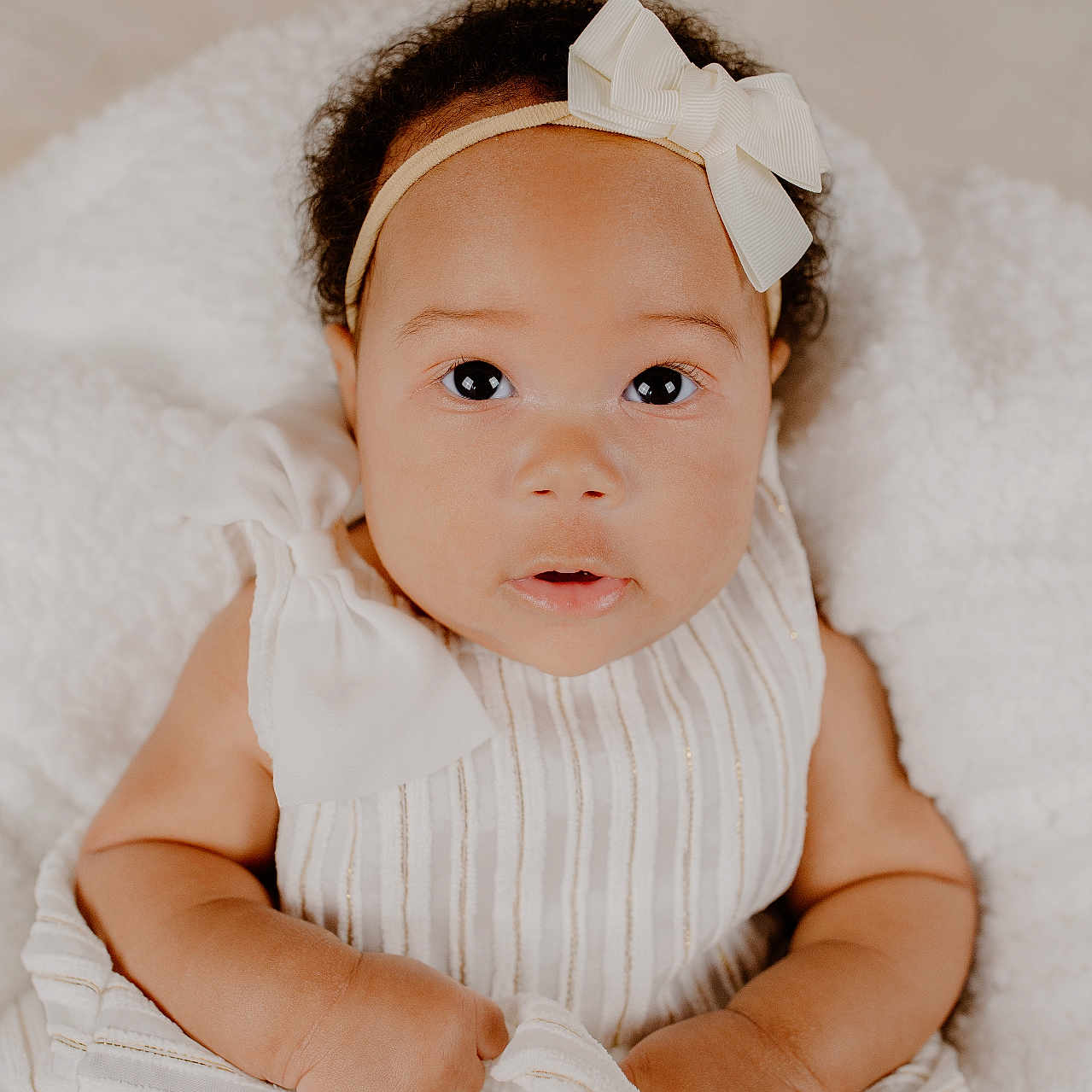 Armiley is registered to the contest to win money with this photo: adorable, baby, bow, child, closeup, curly_hair, cute, expression, gripping, hands, headband, indoors, infant, looking_up, lying_down, portrait, skin, soft_blanket, striped_clothing, white_dress