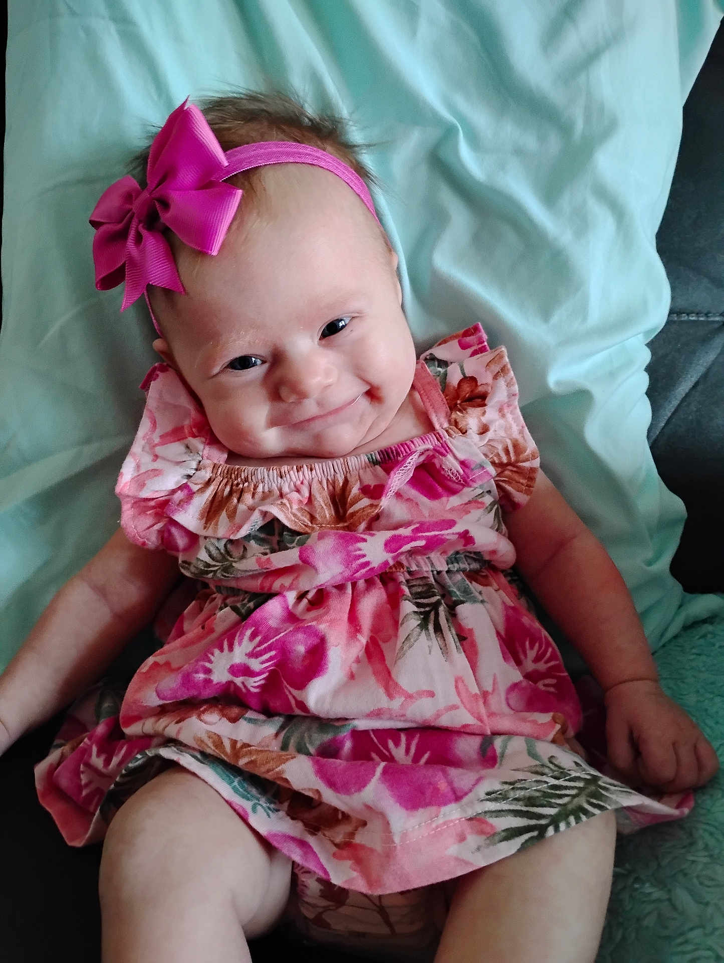 Macie is registered to the contest to win money with this photo: baby, child, smile, pink_bow, headband, dress, floral_pattern, sitting, cute, infant, happy, indoors, skin, arm, leg, fabric, blanket, person, portrait, comfort