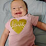 Macie joined the competition — help win amazing prizes! baby, smiling, pink_onesie, gold_heart, love_text, lying_down, bed, happy, infant, cute, skin, face, arm, child, indoors, soft_surface, closeup, joy, expression, toddler