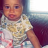 baby, child, infant, sitting, couch, blanket, portrait, face, smiling, tongue_out, blue_eyes, curly_hair, onesie, clothing, indoor, window, blinds, home, close_up, happy