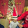 toddler, child, santa_hat, christmas_tree, holiday_decorations, lights, adult_hand, armchair, cozy_room, brick_wall, festive, holiday, indoor, person, clothing, sitting, celebration, home, holding, red_sweater