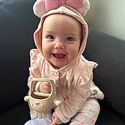 Sophia is registered to the contest to win money with this photo: baby, child, smile, hoodie, pink, bow, toy, sitting, couch, happy, face, cute, indoors, person, infant, clothing, playful, sock, patterned_pants, bright