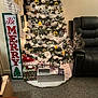 christmas_tree, cat, tabby_cat, presents, wrapped_gifts, leather_recliner, carpet, holiday_decorations, ornaments, garland, indoor, furniture, festive, celebration, cozy, home_interior, sign, wall_art, curious_cat, lights