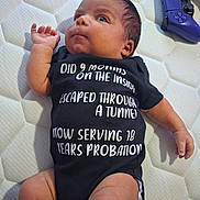 Xavier is registered to the contest to win money with this photo: baby, infant, onesie, text_on_clothing, mattress, bedroom, controller, video_game_controller, purple, cute, lying_down, child, human, skin, hand, foot, expression, clothing, indoors, soft_light