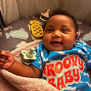 Ryanne is registered to the contest to win money with this photo: baby, smiling, blanket, blue_shirt, groovy_baby, toy, pacifier, pineapple_toy, whale_pattern, cushion, infant, happy, child, closeup, face, cute, soft_texture, indoors, lying_down, colorful_clothing