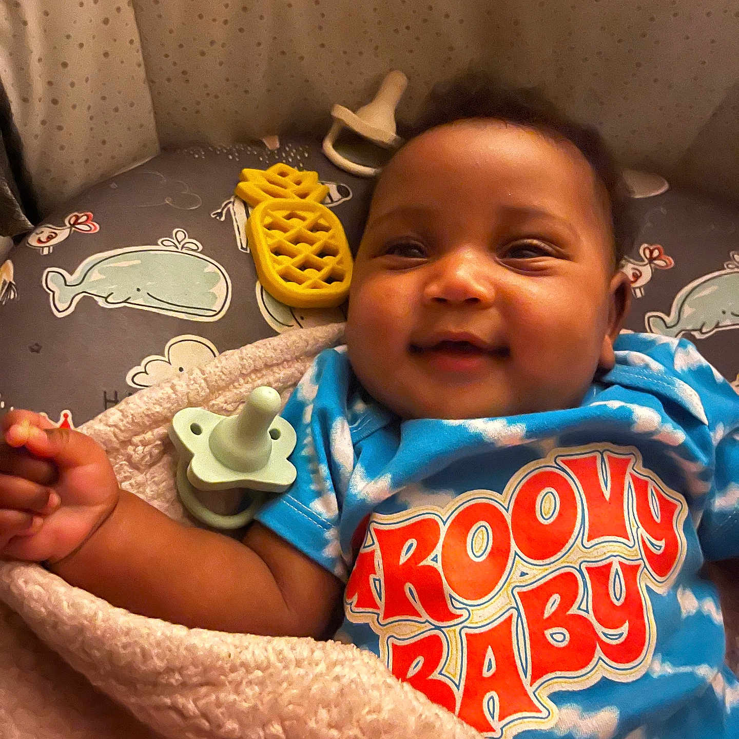 Ryanne is registered to the contest to win money with this photo: baby, blanket, blue_shirt, child, closeup, colorful_clothing, cushion, cute, face, groovy_baby, happy, indoors, infant, lying_down, pacifier, pineapple_toy, smiling, soft_texture, toy, whale_pattern