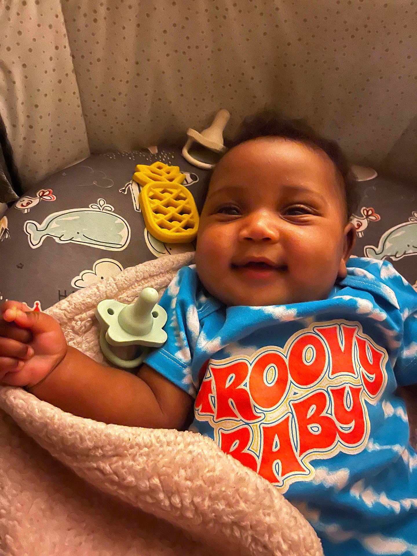 Ryanne is registered to the contest to win money with this photo: baby, smiling, blanket, blue_shirt, groovy_baby, toy, pacifier, pineapple_toy, whale_pattern, cushion, infant, happy, child, closeup, face, cute, soft_texture, indoors, lying_down, colorful_clothing