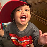 Luca is registered to the contest to win money with this photo: toddler, child, smiling, cap, red_cap, sweatshirt, cartoon, indoor, wooden_furniture, curly_hair, happy, open_mouth, person, clothing, young_child, playful, face, expression, standing, reaching_hand