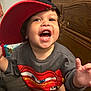 toddler, child, smiling, cap, red_cap, sweatshirt, cartoon, indoor, wooden_furniture, curly_hair, happy, open_mouth, person, clothing, young_child, playful, face, expression, standing, reaching_hand