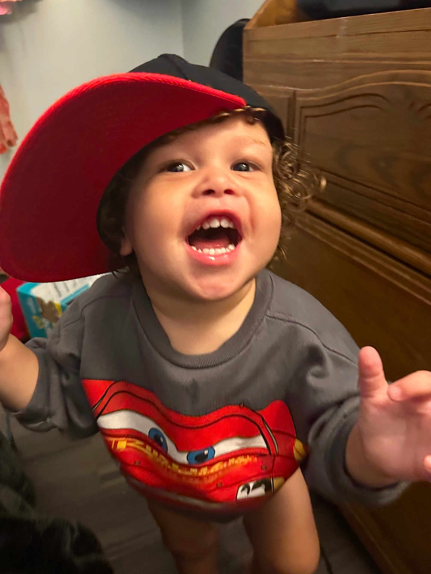 Luca is registered to the contest to win money with this photo: toddler, child, smiling, cap, red_cap, sweatshirt, cartoon, indoor, wooden_furniture, curly_hair, happy, open_mouth, person, clothing, young_child, playful, face, expression, standing, reaching_hand