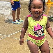 Zoey joined the competition — help win amazing prizes! toddler, child, swimsuit, minnie_mouse, water_play, wet_ground, playground, happy, smiling, curly_hair, yellow_clothing, blue_shorts, crocs, summer, outdoor, sunny, person, fun, splash, play