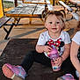 toddler, child, sitting, picnic_table, outdoor, pigtails, minnie_mouse, sippy_cup, pink_shoes, casual_clothing, daylight, bench, wooden_table, playful, cute, smiling, nature, rural, grass, sunlight