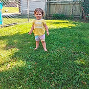 Sapphire is registered to the contest to win money with this photo: toddler, child, grass, lawn, outdoor, fence, gate, yellow_shirt, colorful_shorts, barefoot, smiling, sunlight, daytime, person, happy, young_child, wooden_fence, chain_link_fence, greenery, backyard