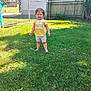 toddler, child, grass, lawn, outdoor, fence, gate, yellow_shirt, colorful_shorts, barefoot, smiling, sunlight, daytime, person, happy, young_child, wooden_fence, chain_link_fence, greenery, backyard