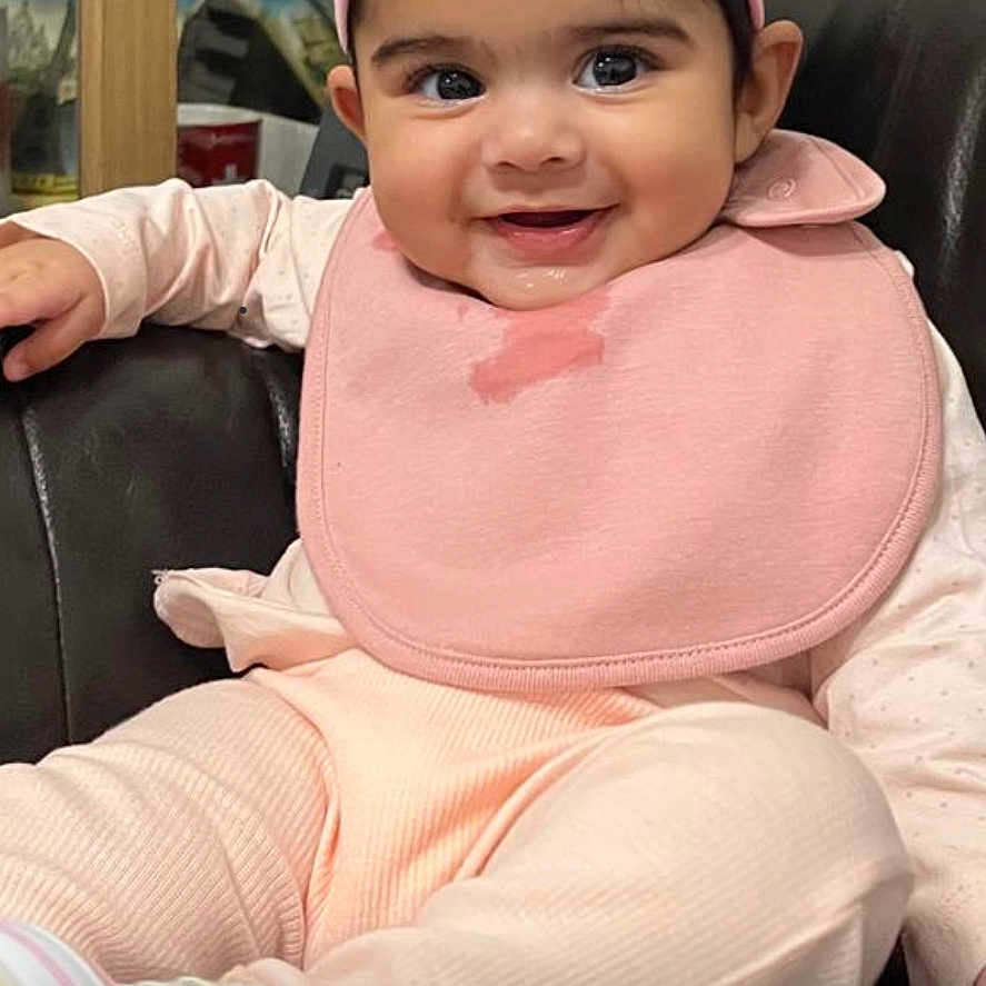 Kinza joined the competition — help win amazing prizes! baby, bib, black_leather, chair, child, closeup, cute, drool, face, flower_accessory, happy, indoor, infant, pastel_clothing, person, pink_headband, portrait, sitting, smiling, young_child