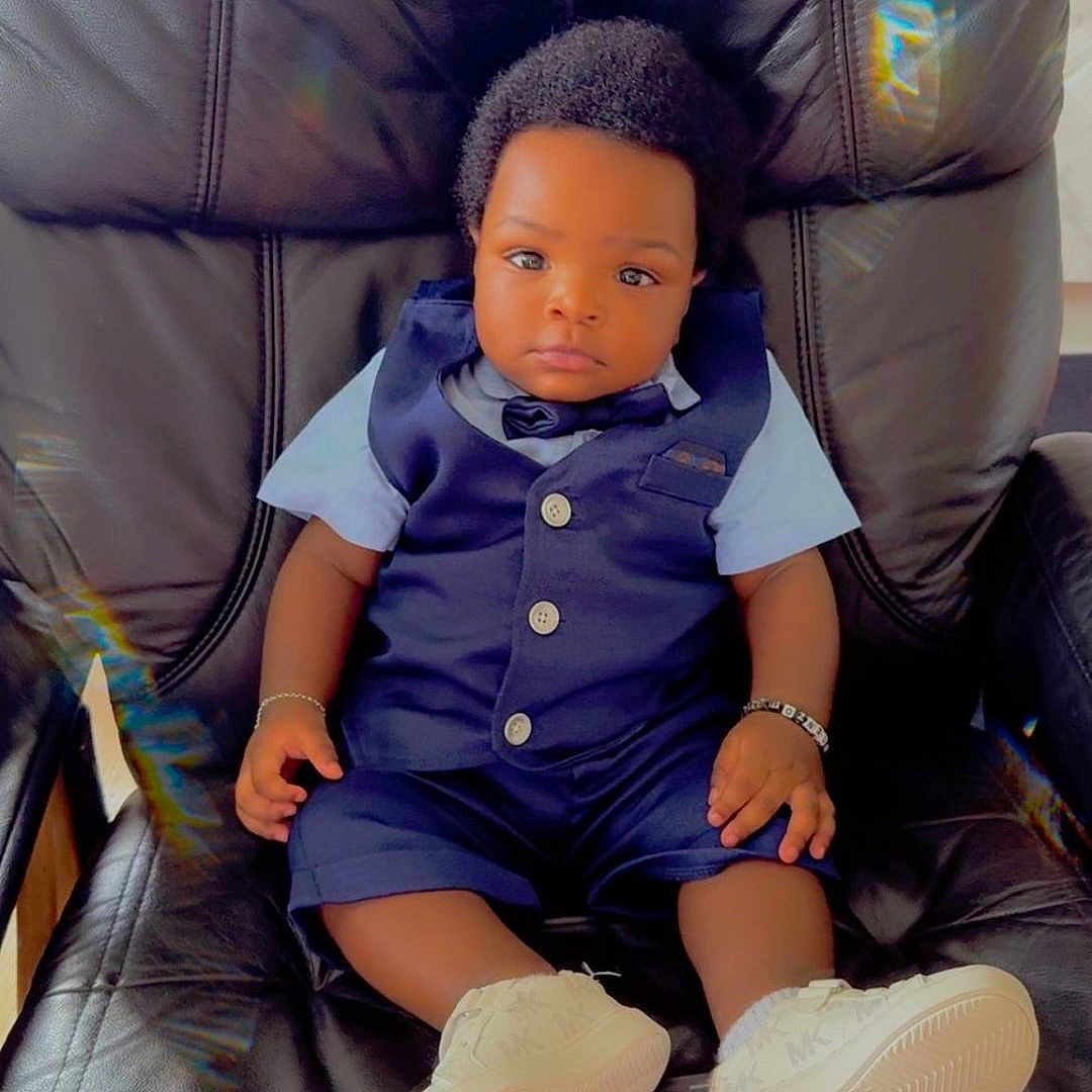 Uriel David a rejoint le concours — aidez-le/la à gagner de superbes lots ! baby, black_hair, bracelet, calm, child, clothing, comfortable, cute, furniture, indoor, infant, leather_chair, looking_at_camera, person, portrait, shorts, sitting, vest, white_shoes, young