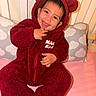 adorable, baby, bear_hood, bed, child, close_up, cloud_pattern, fuzzy_texture, happy, hooded_clothing, indoors, infant, nursery, pillow, portrait, red_outfit, sitting, smiling, tongue_out, toy